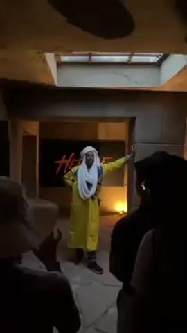 The video shows a group of people inside a room with sandstone walls, standing around a man dressed in a yellow robe, white turban, and blue scarf. He is leaning against one of the walls. A banner with the word "HERITAGE" is visible in the background. A square opening in the ceiling allows some light into the room. The person filming is part of the group and wears a white hat. There are other people in the group whose faces are obscured. The man is addressing the group.