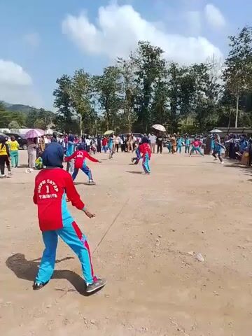 A group of young people in red and blue athletic uniforms are playing a game outdoors in a dusty, open field. The sun is shining brightly, casting shadows on the ground. Some of the players are actively engaged in the game, while others appear to be waiting their turn or observing. Spectators are gathered around the perimeter of the field, some standing and some sitting. Trees and a building are visible in the background. The overall atmosphere appears to be one of a community event or a sporting competition.