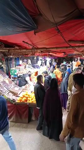 The video shows a crowded marketplace. People are milling around, looking at and buying fruit from stalls. There's a large display of oranges at one stall, and other fruits like dates and pomegranates are visible at another. The market is covered by a makeshift roof of tarps and plastic sheets. The lighting is relatively dim. People are dressed in a variety of clothing, from casual wear to more traditional attire. There's a general sense of activity and commerce.