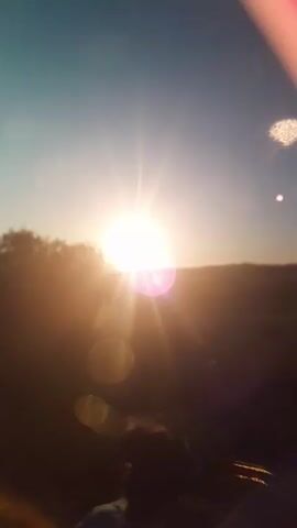 A vertically oriented, low-resolution video captures a sunset. The sun is a bright, white orb with visible rays and lens flares, casting a warm orange glow across the scene. Silhouetted against the sun is a distant treeline. In the foreground, a person's dark hair and shoulders are visible from behind. The sky transitions from a pale blue at the top to a warm orange near the horizon. The overall quality of the video is blurry and grainy, with noticeable lens artifacts such as circular light refractions and a pinkish hue on the left side of the frame, possibly from the camera lens or a filter. The sun appears to be setting behind a hill or elevated terrain. The video seems to be taken outdoors in a natural setting.