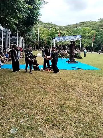 Here’s a plain text description of the video:

The video depicts a grassy area where a group of people are gathered around a blue mat. Several individuals, dressed in black shirts and pants, stand near the mat. Two other people are sitting on the mat. The setting appears to be an outdoor event, with spectators seated on the ground and standing in the background. A tent-like structure is visible in the distance, also on the blue mat. Trees and hills are visible in the background, suggesting the event takes place in a park or open space. One person on the mat appears to be opening a box. A few people seem to be observing the activity on the blue mat. Overall, the scene suggests a ceremony or performance is about to begin.