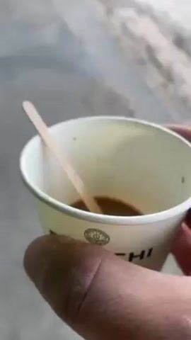 The video shows a hand holding a white paper cup with a small amount of dark liquid inside, likely coffee. A wooden stir stick is in the cup. The camera pans up to show a view of a street, sidewalk, and a building across the street. In the distance, a Bulgarian flag is visible next to a monument. The environment appears to be a small town square on a clear day.