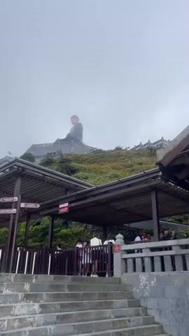 The video pans across a cloudy, mountainous landscape. In the background, a large, dark statue of a seated Buddha is visible on a hilltop. In the foreground, there are covered structures with wooden roofs and railings, and a set of concrete stairs leading upwards. Several people are standing around and walking through these structures. Signposts with text in a foreign language are also visible. The weather appears overcast or foggy.