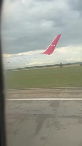 The video shows a view from an airplane window. The wing of the plane, painted red with a white logo, is visible in the foreground. Outside the window, a grassy field stretches towards a tree line under a cloudy sky. A runway with markings is visible at the bottom of the frame. The plane appears to be on the ground, possibly taxiing or preparing for takeoff.