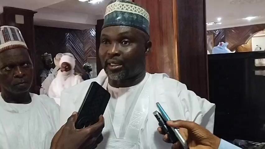 The video features a man with a dark complexion, a salt-and-pepper beard, and a dark blue and white striped hat, being interviewed by two people holding smartphones with microphones pointed towards him. He is wearing a white traditional outfit. The interview is taking place indoors, with several other people in traditional white clothing and hats visible in the background. There is a dark wooden cabinet behind the main subject, and the room has a subdued, warm lighting. The focus is tightly on the man being interviewed, with the smartphones and hands of the interviewers prominent in the foreground. The overall setting suggests a formal or official environment.