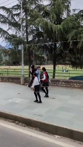 This video captures a street scene with a sidewalk and a road. On the sidewalk, there are several people walking and standing. A young woman in a white shirt and black skirt walks to the left, while a person in a black jacket and jeans walks to the right, talking on a cell phone. Further down the sidewalk, a group of people are walking together. In the background, there is a park or open field with a green fence. Several trees are visible, including large evergreen trees. Mountains form the backdrop under a cloudy sky. A utility pole with wires and a sign is prominent in the foreground. A man is sitting on a park bench. The overall scene suggests a public space in a hilly or mountainous area.