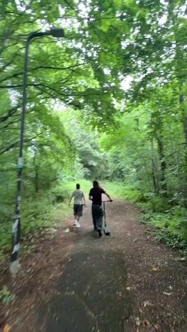 Two people are walking down a dirt path surrounded by green trees. The person in the front is walking with a scooter. The sky is overcast.