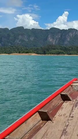 A close-up perspective from a wooden boat shows the turquoise water and forested mountains in the distance under a partly cloudy blue sky. The boat's weathered wooden deck and red trim are in the foreground.