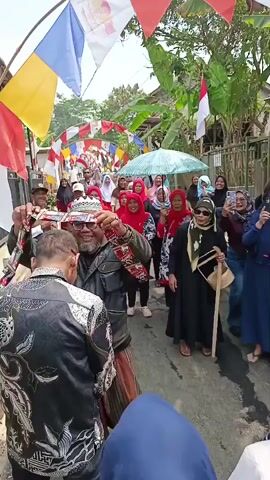 The video captures an outdoor scene where a group of people are gathered on a narrow street. Several colorful flags and pennants are strung across the street, creating an arched effect. In the foreground, two men are interacting. One man, wearing a black jacket with a patterned design and a white cap, is shaking hands with another man. The second man is wearing a dark jacket and a striped sarong. Behind them, a crowd of people, mostly women wearing headscarves, are watching and some are holding up their phones, presumably to record the event. One woman in the middle of the crowd is holding a light blue umbrella. Indonesian flags are also visible in the background. The environment appears to be a residential area with houses lining the street. The overall atmosphere seems festive.