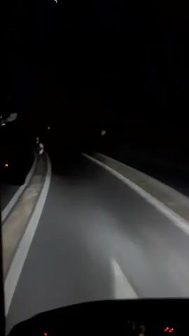 A first-person perspective shot from inside a vehicle at night shows a dark road with headlights illuminating the path ahead. The road has white lane markings and a guardrail on the left. Traffic signs are visible along the roadside. The vehicle's dashboard lights are also dimly lit.
