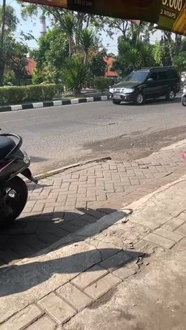 A busy road is captured from a sidewalk. Several motorcycles and a few cars are seen driving on the asphalt road. The motorcycles are predominantly driven by individuals wearing helmets and the riders appear to be of average build. One motorcycle has two people on it, with the passenger sitting behind the driver. The environment appears to be an urban or suburban setting with trees and some buildings visible in the background. The weather seems to be sunny, casting shadows on the sidewalk.