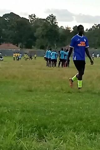 The video shows a soccer field with two teams of players. One team is wearing light blue jerseys and black shorts, while the other team is wearing yellow jerseys and black shorts. The light blue team is gathered in a group in the center of the field, while the yellow team is spread out. A player in a light blue jersey and black shorts walks along the right edge of the field. The field is green and grassy, and there are trees in the background. The sky is cloudy. The players appear to be standing or walking.