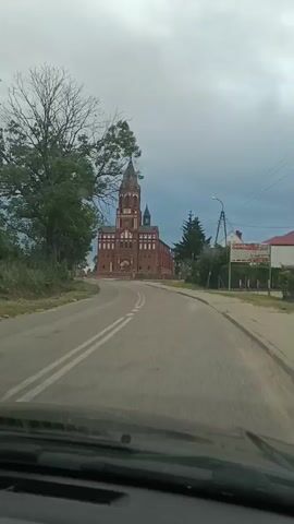 The video begins with a view from inside a vehicle looking out at a road leading towards a large, brick church with a tall, spired bell tower. The church has a prominent facade with arched windows and detailing. Trees are visible on the left side of the road, and to the right, there is a utility pole and some buildings. The sky is overcast. The vehicle appears to be moving forward on the asphalt road, which has double yellow lines in the center. The camera is positioned at dashboard height. The video is short, showing the church getting slightly closer as the vehicle drives towards it.