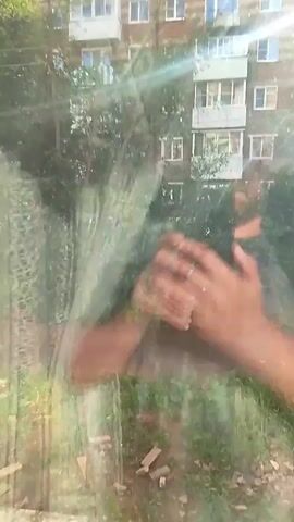 This video appears to be filmed from indoors, looking out through a window. The primary subjects are hands, visible in the foreground and blurred by the camera's focus and possibly smudges on the window. Beyond the hands, a building with multiple floors and balconies is visible, appearing to be an apartment building. Trees and foliage are in front of the building, obscuring parts of it. The bright sunlight suggests it is daytime, possibly morning or afternoon, and the sun's glare is significant. The overall scene suggests an urban or suburban residential area in Prisel'e, Russia, with a focus on the view from a window. The activity seems to be passive observation, with the hands potentially interacting with the window or the view. There is no discernible text in the video. The quality of the footage is low, with significant blur and lens flare.