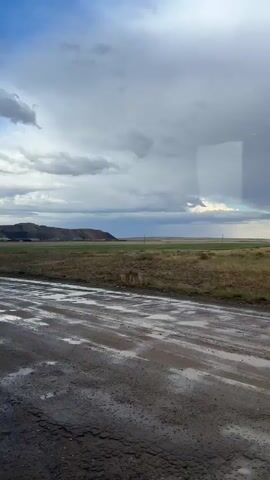 This is a first-person perspective video of a person traveling on a muddy dirt road. The road is wet and filled with puddles, suggesting it has recently rained. The environment is rural, with fields of dry grass and a distant hill under a cloudy sky. The camera is angled down towards the road, with the sky and distant landscape visible in the background. The overall impression is one of a damp and overcast day in a rural setting.