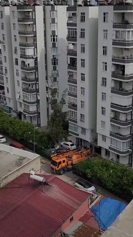 An aerial view shows a street between two tall apartment buildings. An orange truck with a large tank, likely a septic truck, is parked on the street. There are several parked cars and some trees and bushes lining the street. In the foreground, the rooftops of two buildings are visible, one with a red metal roof and another with a blue tarp. The overall impression is of an urban residential area.