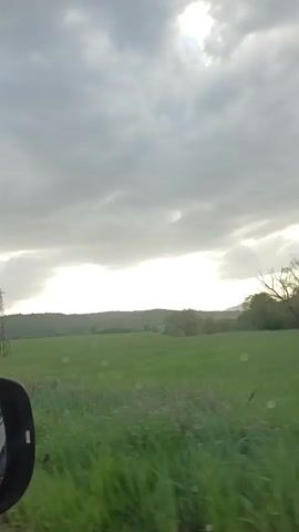 A car drives down a road through a rural area. The sky is cloudy with patches of sunlight. The land on either side of the road is mostly green fields and forests. An electrical tower is visible in one of the fields. Rain is on the windshield of the car. There are other cars visible on the road ahead. The side mirror of the car is visible in the lower left of the frame in some shots.