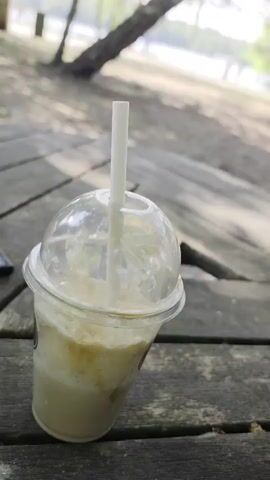 The video shows a disposable cup of milky liquid with a straw, sitting on a weathered wooden picnic table. Beyond the table, the scene opens to a grassy area with trees, leading to a body of water in the background. Two figures can be seen standing near the water's edge. Sunlight filters through the trees, casting shadows on the ground and the table. The camera angle is low, emphasizing the cup in the foreground and the natural setting beyond.