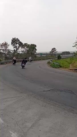 The video shows a winding paved road with a slight incline. Several motorcycles and a bicycle are visible on the road. The environment appears to be a rural or mountainous area with trees and vegetation lining the sides of the road. The sky is overcast, suggesting a cloudy day. The road surface shows signs of wear and tear, with cracks and patches visible. There are guardrails along some sections of the road, and a bridge-like structure is visible in the background. The overall impression is one of an outdoor scene in a somewhat undeveloped area.