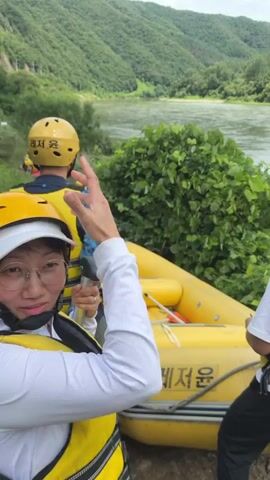 The video begins with a close-up shot of a person wearing a white visor, glasses, and a yellow life vest. Behind them, another person in a yellow helmet and yellow life vest is visible, facing away from the camera. To the right, the edge of a yellow raft with the word "레져" (Leisure) in Korean lettering is prominent. The raft appears to be on the shore of a river. The background reveals a wide, calm river flowing between lush green, tree-covered hills under a partly cloudy sky. As the video progresses, the camera pans slightly, showcasing more of the river and the mountainous landscape. Other people, also wearing yellow life vests and helmets, are seen gathered near other rafts further down the riverbank. The overall atmosphere suggests preparation for or the beginning of a rafting activity. The camera continues to pan, offering a wider view of the natural scenery, with the river reflecting the sky and the dense greenery of the hills dominating the horizon. The video concludes with a sweeping view of the river and the surrounding forested mountains.