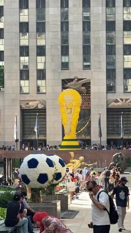 The video shows a large yellow trophy replica, possibly made of Legos, standing in front of a tall building with many windows. In the foreground, there is a giant soccer ball sculpture decorated with white and blue floral elements. A crowd of people is gathered around the sculptures and in front of the building. Some people are taking pictures, while others are walking or standing. A golden statue of a figure in motion is visible behind the soccer ball sculpture. The overall atmosphere appears to be festive and public.