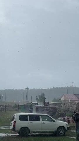 The video shows a white minivan parked in a grassy area. There are several structures in the background, including a wooden fence, a building with a red roof, and a large metal tank. A tractor is partially visible behind the minivan. The sky is overcast, and it appears to be raining or snowing. There are numerous white specks falling from the sky, which are likely precipitation. The overall environment appears to be rural.