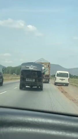 The video shows a highway scene from the interior of a moving vehicle. Ahead, in the same lane, a black van is transporting a large, wooden structure. To the right, a white minivan is visible on the shoulder of the road. The sky is partly cloudy, and a hilly landscape is visible in the distance.