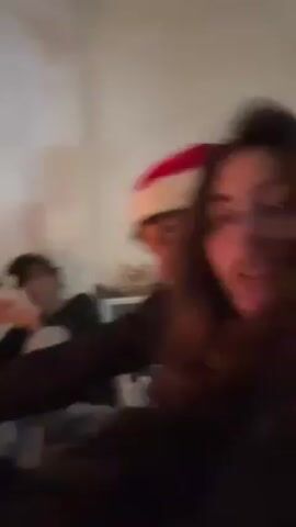 A low-lit video shows a young woman holding the camera, angled towards herself and a young man behind her. The man is wearing a Santa hat. A third person sits on the floor in the background to the left. Behind the couple, a bookshelf is visible with numerous books. The couple appears to be lip-syncing or singing along to a song. The camera angle changes several times throughout the video, shifting focus between the faces of the woman and the man in the Santa hat. The overall setting appears to be indoors, likely a home.