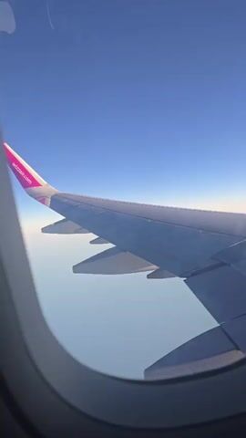 A first-person perspective video shows an airplane wing and the sky through an airplane window. The wing is white with pink accents near the tip, featuring the Wizz Air logo. The sky is a clear, bright blue, with a faint orange glow near the horizon, suggesting it is either sunrise or sunset. Clouds are not visible. The video appears to be taken while the plane is in flight.