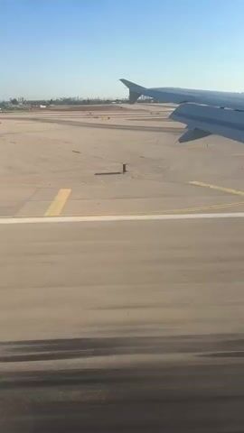 This media file captures the view from an airplane window as it takes off from an airport. The primary subjects are the airplane's wing and the runway. The scene is an airport tarmac with a clear blue sky above, suggesting it is daytime.

The activity taking place is an airplane takeoff. We see the runway markings and the vast expanse of the airport grounds. In the distance, a cityscape can be seen, hinting at the urban environment surrounding the airport. The wing of the plane is prominently featured in the foreground, angled upwards, indicating the aircraft is ascending. The runway surface shows tire marks, evidence of previous landings and takeoffs.

Notable details include the clear weather and the bright daylight, indicated by the strong shadows cast by the plane's wing and other airport structures. The location cues provided are "Sdot Dan Regional Council, Israel," which places the airport in that region. There is no visible text in the media file. The overall impression conveyed is one of motion and departure, with a sense of ascent and transition.