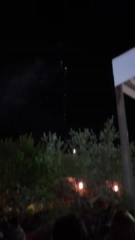 A fireworks display takes place at night. Fireworks explode in the sky, emitting pink and purple light. The ground below is dark, with silhouettes of trees visible. A structure with a white roof is partially visible on the right. Further down, a table with a white cloth is set up, with a cake and illuminated letters that spell out "1BR." The scene is lit by ambient light and the glow of the fireworks and decorations.