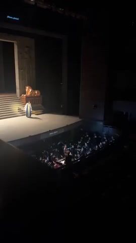 The video shows a man on a stage in what appears to be a theater. The stage has a backdrop resembling an ancient Egyptian setting, including a staircase and what looks like a golden lion statue. The man is wearing a long, light-colored robe with blue trim. He is standing on the stage and appears to be gesturing with his hands. Below the stage, in the orchestra pit, is a group of musicians playing instruments. The audience is seated in the darkness beyond. The lighting is focused on the stage, making it the brightest area in the frame.