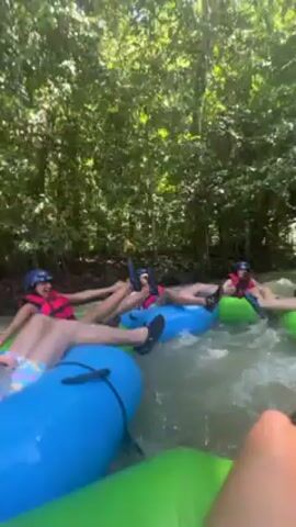 The video shows several people river tubing. They are wearing life vests and helmets. The tubes are various colors including blue and green. They are floating down a river that is surrounded by dense green foliage. The camera appears to be held by someone also in a tube.