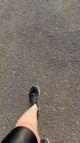This media shows a first-person perspective of someone walking on a paved path. The path is marked with bicycle symbols, indicating it's a bike path or a shared recreational trail. The scene opens with a close-up of the walker's feet in athletic shoes and black leggings, stepping on the coarse, grey asphalt. As the walk progresses, the camera pans up, revealing the surrounding environment.

The setting appears to be a park or a recreational area near a body of water, possibly a lake or a large river, given the expansive view of water and distant tree line under a bright, sunny sky. The time of day is clearly morning or early afternoon, as indicated by the low angle of the sun and the long shadows, creating a pleasant, bright atmosphere. The weather seems fair, with a clear blue sky and a few scattered clouds.

Notable details include a metal fence to the left of the path, and in the distance, there are some park amenities such as benches or signs and a trash can. The overall mood is peaceful and suggests a leisurely activity like a walk or a run in a serene natural setting. The location is identified as Падлабенскі сельскі Савет, Belarus, providing a specific geographical context for this outdoor scene. There is no visible text in the media, other than the bicycle symbols on the path.
