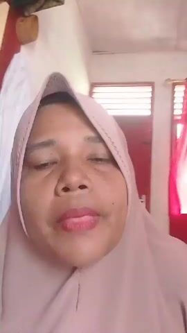 A person wearing a light pink hijab is facing the camera, their mouth is open as if they are speaking. They have brown eyes and their lips are colored with a light pink lipstick. Behind them is a room. A window is visible with sheer red curtains. A red door can also be seen.