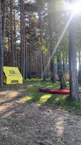 The video shows a campsite scene. In the foreground, there is a dirt path with scattered pine needles. To the left, a yellow tent is set up. In the middle ground, a red inflatable kayak lies on the grass. Several tall trees, primarily pines and birches, are visible throughout the scene, with sunlight filtering through the leaves. Further back, there are glimpses of wooden structures and a lake or body of water. A blue and yellow patterned blanket or tarp is draped over a low structure on the right side of the frame, near a trailer-mounted wooden structure that appears to be a sauna or small building with a chimney. The overall environment suggests a natural, outdoor recreational area. There are no people visible in the video.