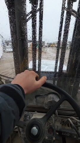 The video shows the view from inside the cab of a forklift on a rainy day. A hand is visible on the steering wheel. The view through the windshield shows a wet, muddy yard scattered with wooden debris, pallets, and some industrial buildings in the background.