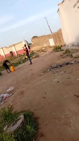 The video depicts two individuals in an open, seemingly undeveloped area littered with trash. One individual, wearing dark clothing, is bent over, handling a yellow jerrycan. The second individual, dressed in a red shirt and jeans, stands watching. The setting appears to be an outdoor, dirt-covered area with buildings in the background, one of which has a visible door. The sky is visible, suggesting it is daytime.