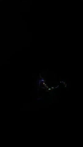 The video begins with a close-up shot of multi-colored lights strung together, then pans to a dark outdoor scene. Silhouettes of several people are visible. Some individuals are standing, while others appear to be sitting or bending over. In the background, more lights, some red and some white, are intermittently visible. The overall impression is of a gathering or event taking place in the dark.