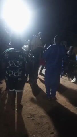 The video shows a group of people gathered outdoors at night. A bright light source illuminates the scene, making it difficult to discern details. The group consists of both men and women of varying heights. Some are wearing casual clothing, while one man is wearing a blue robe. They are standing on a dirt surface and appear to be watching something, potentially a performance or event. In the background, there are chairs and additional people. The lighting makes it hard to see any specific objects clearly. The atmosphere appears to be festive, with music playing in the background.