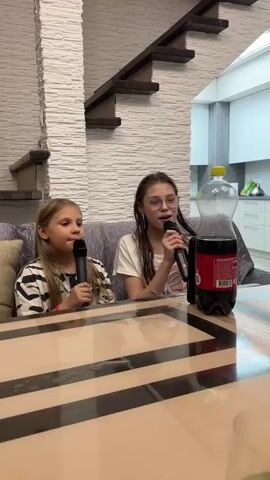 Two young girls are singing karaoke. A girl with glasses holds a microphone with one hand, and she sings into it. The other girl is also holding a microphone and singing. Both are looking forward at the camera. They are seated on a gray couch that is partially covered with pillows. In front of them is a shiny, striped table. On the table is a glass water bottle balanced atop a plastic bottle of Coca-Cola. Behind them is a white wall with bricks, and a wooden staircase with an ornate bannister. In the background is a kitchen.