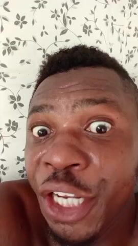 The video features a close-up shot of a Black man's face. He has a short, dark, curly haircut. His skin appears sweaty or moist. His eyes are wide and focused. He is smiling intensely, revealing his white teeth. The background is a white wall covered with a floral pattern of gray and black. The man appears to be speaking, and he is looking directly into the camera.