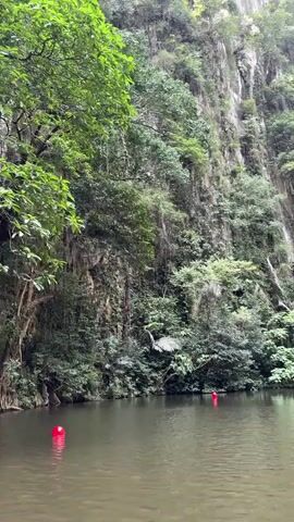 A person is visible in the distance, near a sheer rock face covered in vegetation. The person appears to be wearing red clothing. The person is standing in the water, which is calm and reflects the surrounding cliffs. There are two red buoys in the water, one closer to the viewer and one further away. The environment is lush and tropical, with dense foliage growing on the cliffs. The scene appears to be a natural landscape, possibly a lake or river surrounded by cliffs.