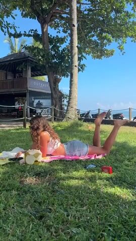 A young woman is lying prone on a pink towel in a grassy area. She has long, curly brown hair. She is wearing a white halter top and denim shorts. To her right, there is a partially consumed coconut and a small, red JBL speaker. The woman appears to be looking at or using a tablet or electronic device. Behind her, there is a wooden structure with the word "OUTKAST" visible on its sign. Beyond that, there is a view of the ocean under a bright blue sky. Several motorbikes are parked along a road in the background. The sun is shining brightly, and the scene suggests a tropical or coastal location.