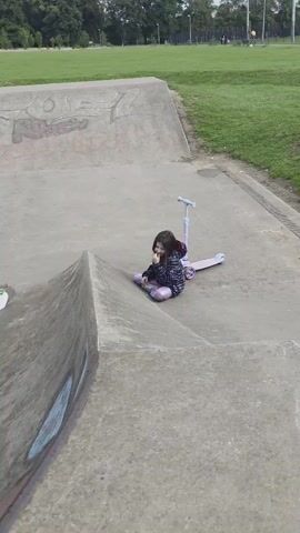 A young girl sits cross-legged on the concrete edge of a skate park, her pink scooter resting behind her. She is wearing a dark jacket with a patterned design and light-colored pants. The girl appears to be touching her face or mouth with her right hand. The skate park is empty except for her, and the surrounding area is a grassy field with trees and some distant structures. The sky is overcast.