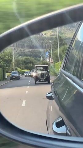 The video is captured from the perspective of someone in a moving car, looking into the side-view mirror. The mirror reflects a road with other vehicles. Directly ahead in the reflection is a vintage car, possibly from the early 20th century, with a dark body and a light-colored top. Its headlights are on. Behind the vintage car, two other cars are visible on the road. The road is paved, with white lane markings. On either side of the road, there are hedges and some trees. In the background, there is a hill covered with trees. The sky is visible above the hill. The overall impression is of a sunny day, with the vehicles driving on a suburban or rural road.