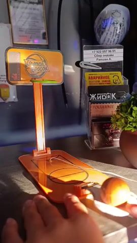 The video shows a close-up shot of a mini basketball hoop game on a dark surface, possibly a desk. An orange backboard with a logo that reads "Крутой бросок" (Cool shot) is attached to an orange pole, which is mounted on an orange base shaped like a basketball court. A small orange basketball is connected to the base by a string and is positioned near the bottom of the frame. A hand, partially visible, is about to press a button or lever on the right side of the base. The background is cluttered with various items, including business cards, a small plant, and what appear to be official documents or signs. The lighting suggests it might be daytime, with sunlight casting shadows on the objects. The video captures the action of a player interacting with the game.