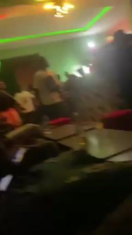 The video shows a dimly lit room, possibly a bar or nightclub, with several people present. The lighting changes between shades of red, blue, green, and purple, creating a party atmosphere.  People are seen sitting in cushioned chairs around tables. There are also people moving around in the background. The video seems to be taken from a seated position.  Glasses and bottles are visible on the tables. The specific activities of the people are unclear due to the low lighting.  Music is playing in the background.