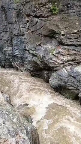 The video shows a turbulent, muddy river flowing through a narrow canyon. The canyon walls are steep and rocky, with some vegetation growing at the top. The water appears to be moving quickly and is churning with white foam. There are no people or animals visible in the video. The overall impression is one of natural power and rugged beauty.