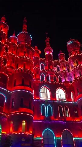 This video shows a night view of a castle-like building illuminated with red, pink, and blue lights. The building has multiple towers with pointed roofs, arched windows, and decorative elements. The lights are strung along the edges of the structure, outlining its architectural features. The background is dark, indicating nighttime.